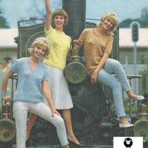 Puede incluir: Tres mujeres con diferentes suéteres de punto posan frente a un tren vintage. La mujer de la izquierda lleva un suéter azul claro, la mujer del medio lleva un suéter amarillo y la mujer de la derecha lleva un suéter beige. Los suéteres son todos de manga corta y tienen un patrón de punto de cable. Las mujeres están de pie frente a una gran locomotora negra. La locomotora tiene una gran rueda redonda y una pequeña ventana redonda. Las mujeres sonríen y miran a la cámara. La imagen es en color y tiene un aspecto vintage.