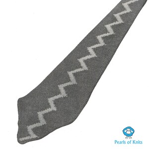 May include: A gray knitted scarf with a white zig-zag pattern. The scarf is long and narrow, and the pattern is repeated throughout the length of the scarf.