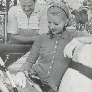 May include: A woman wearing a gray knitted cardigan with a button-up front and a white scarf. She is sitting in a car with a cat on her lap. The woman is wearing white gloves and a headband. The car has a black leather interior.