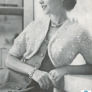 Pode incluir: Um bolero branco de crochê com uma borda festoneada e mangas curtas. O bolero é usado sobre um vestido preto com botões.