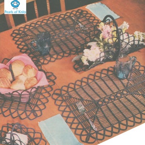 Pode incluir: Três tapetes de crochê de cor castanho escuro com forma quadrada e uma borda decorativa. Os tapetes estão sobre uma mesa de madeira com uma toalha azul e uma toalha rosa. Há um copo de água e um garfo em um dos tapetes.