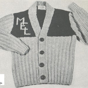 May include: A grey knitted cardigan with a black and white "MET" design on the front. The cardigan has a V-neck and five black buttons.