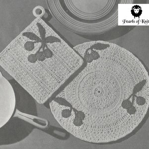 Puede incluir: Dos agarraderas de olla de ganchillo con un diseño de cerezas. Las agarraderas son blancas con cerezas y hojas de color gris oscuro. Las agarraderas están sobre un fondo gris. Pearls of Knits.
