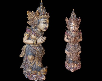 Op de afbeelding: Twee ingewikkeld gesneden houten beelden van Balinese godheden, beide met sierlijke hoofdtooien en gouden accenten. De beelden staan in een gebedshouding met gevouwen handen.