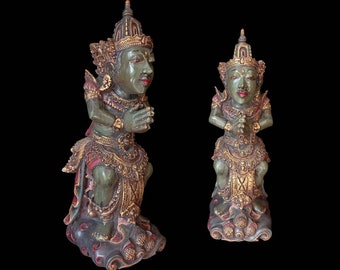 Op de afbeelding: Twee groene en gouden houten beelden van figuren in een knielende houding met gevouwen handen in gebed. De beelden zijn ingewikkeld gesneden en hebben sierlijke details.
