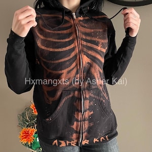 May include: Black zip-up hoodie with a brown skeleton rib cage design. The hoodie has a drawstring hood, front pockets, and the text "Hxmangxts (By Asher Kai)". The design includes paint splatter details.