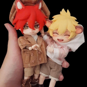 Op de afbeelding: Twee beweegbare poppen in anime-stijl. De linkerpop heeft felrood haar, vossenoren en draagt een bruine geruite baret, een bruin jasje met dubbele rij knopen, beige shorts en bruine veterschoenen. De rechterpop heeft blond haar, ronde oren en draagt een roze hoodie, beige shorts en zwarte schoenen.