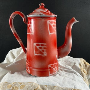 Peut inclure: Cafetière en émail rouge avec un motif géométrique blanc. La cafetière a une poignée et un bec verseur. Elle est posée sur un tissu blanc avec une bordure dorée.