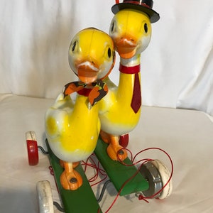 Op de afbeelding: Een vintage trek speelgoed met twee gele eenden met zwarte ogen en rode snavels. De eenden staan op een groen houten platform met rode en witte wielen. De eend achterin draagt een zwarte hoed en een rode stropdas.