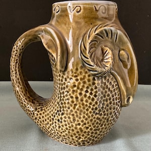 Könnte beinhalten: Eine braune Keramiktasse in Form eines Widderkopfes mit einer strukturierten Oberfläche. Die Tasse hat einen Griff, der wie der Hals und die Hörner des Widders geformt ist.