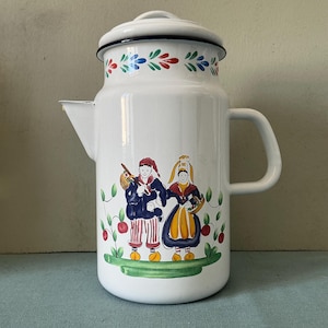 May include: White enamelware coffee pot with a lid and handle. The pot features a colorful floral design around the rim and a painted scene of two figures in traditional clothing. The figures are standing on a green base with red berries.