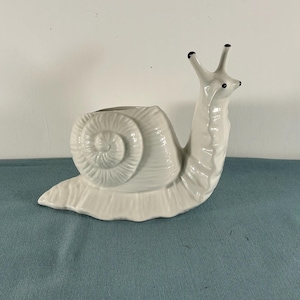 May include: A white ceramic snail-shaped planter. The snail has a detailed shell, a textured body, and two antennae with black tips. The planter's opening is on the snail's back, and it sits on a flat base. The overall design is whimsical and decorative.