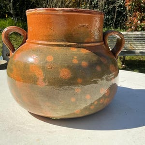 May include: A large, round, ceramic pot with two handles. The pot is glazed in shades of brown and green, with orange polka dots. The pot sits on a white surface, with a bench and trees in the background. This decorative pot is suitable for indoor or outdoor use.