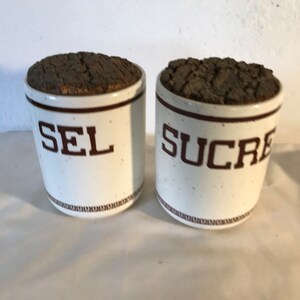 May include: Two white ceramic canisters with cork lids. The canisters have brown trim and the words "SEL" and "SUCRE" printed on the front.