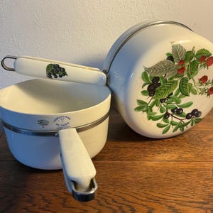 May include: Two white porcelain cooking pots with metal rims and handles. One pot has a lid and a painted design of berries and leaves. The other pot has a handle with a berry design. The pots are on a wooden surface.