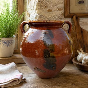 May include: A large, rustic, glazed ceramic pot with two handles. The pot features a rich, earthy color palette of brown, red, and black. It sits on a wooden surface, suggesting a kitchen or pantry setting. The pot's design evokes a sense of traditional craftsmanship.
