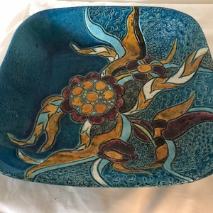 Puede incluir: Un plato cuadrado de cerámica con fondo azul y un diseño floral en tonos de amarillo, marrón, morado y blanco.