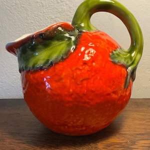 Op de afbeelding: Een keramische kan met een helderrode body, geaccentueerd met groene bladvormige details en een gebogen groene handgreep. De kan heeft een glanzende afwerking en een witte rand, perfect voor het serveren van drankjes. Ongeveer 20 cm hoog.