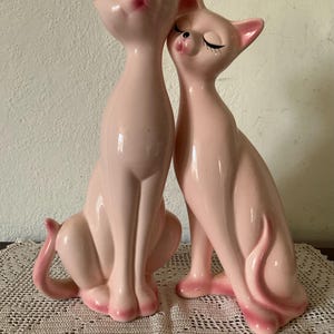 Peut inclure: Deux grandes figurines de chat en céramique rose clair, brillantes, côte à côte. Le chat de gauche a les yeux grands ouverts, tandis que celui de droite a les yeux fermés. Ils ont des oreilles pointues, de longs cous et des accents rose foncé.