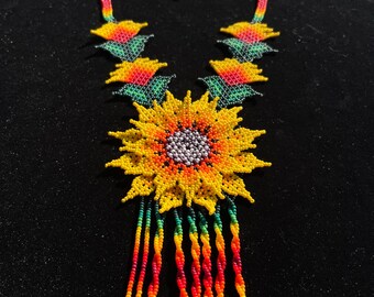May include: A beaded necklace with a sunflower design. The necklace is made of colorful beads in shades of yellow, orange, red, green, and black. The sunflower is the centerpiece of the necklace and has a detailed design with a yellow center and brown petals.