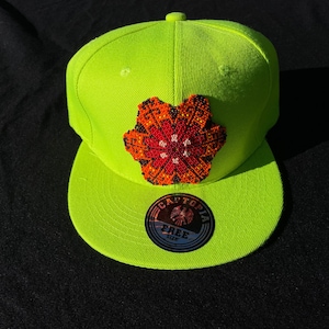 May include: A neon green flat-brimmed baseball cap with a beaded floral design in red, orange, and white. The cap features a circular sticker with the text "CAPTOPIA" and "FREE SIZE".