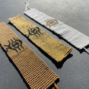 May include: Three beaded bracelets with a geometric design. The bracelets are in gold, white, and black.