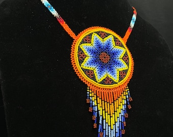 May include: A beaded necklace with a large, round pendant featuring a colorful starburst design. The pendant is surrounded by a fringe of beads in shades of orange, yellow, and blue.