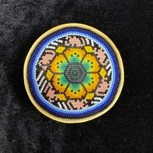 May include: A round, wooden coaster with a colorful beaded design. The center of the design features a flower with yellow, orange, green, and blue beads. The outer edge of the coaster is surrounded by a blue beaded border.