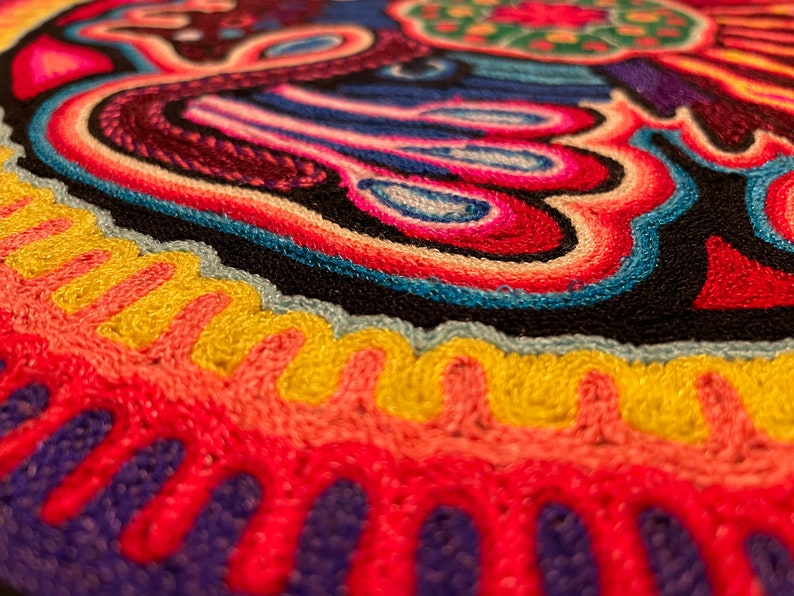 May include: Close-up of a colourful embroidered textile with a circular design. The design features a variety of colours, including red, blue, yellow, green, and white. The textile is made of a soft, textured fabric.