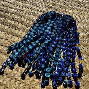 May include: A collection of blue and white beaded necklaces with black string. The necklaces are arranged in a fan shape on a woven mat.