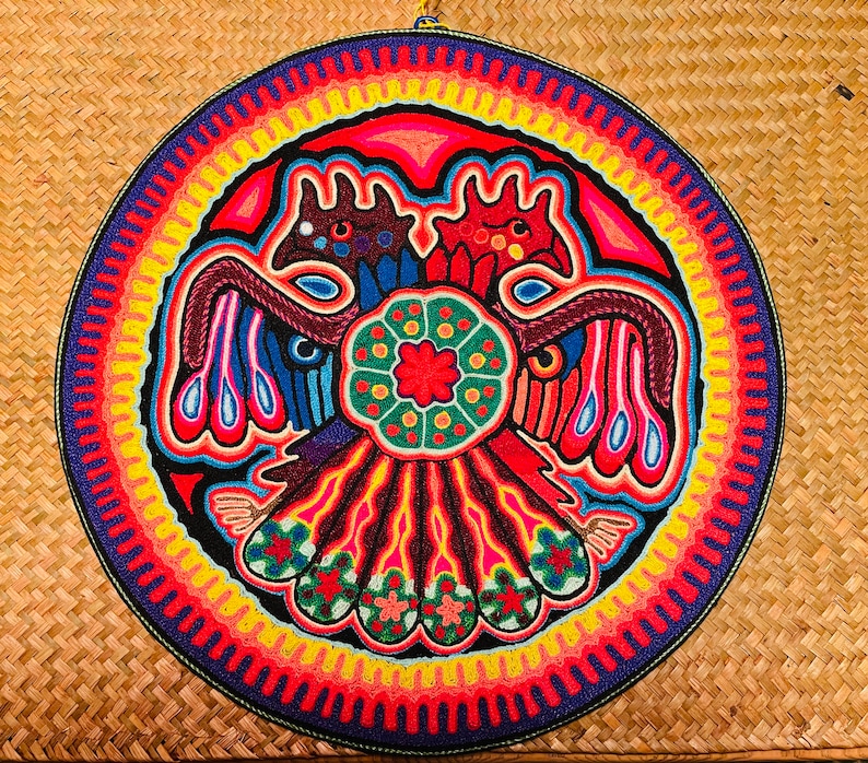 May include: A colourful circular wall hanging with a beaded design of two stylised birds with a central flower motif. The background is a series of concentric circles in shades of red, yellow, and blue.