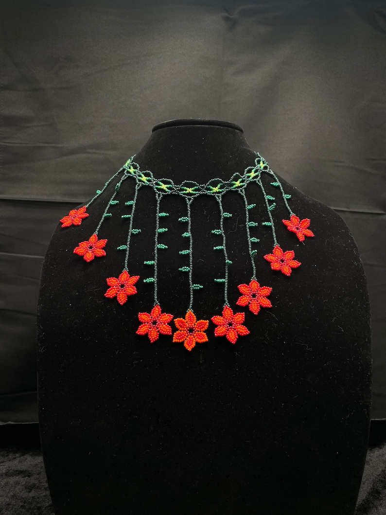 May include: A beaded necklace with red flowers and green leaves. The necklace is made of small beads and is strung together in a delicate pattern. The flowers are arranged in a cascading design, and the leaves are interspersed throughout the necklace.