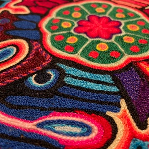 May include: Close-up of a colourful embroidered fabric with a central circular design featuring a pink flower with green and yellow accents. The fabric is made of a variety of colours, including red, blue, green, yellow, and white.