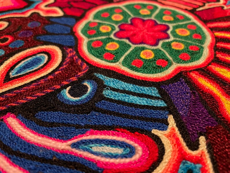 May include: Close-up of a colourful embroidered fabric with a central circular design featuring a pink flower with green and yellow accents. The fabric is made of a variety of colours, including red, blue, green, yellow, and white.