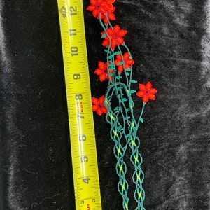 May include: A beaded flower garland with red flowers and green leaves. The garland is approximately 12 inches long.