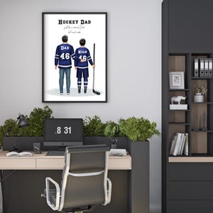 Pu&ograve; includere: Stampa incorniciata con padre e figlio in maglie da hockey, con la scritta "Hockey Dad". Le maglie blu hanno dettagli bianchi e il numero 46. L'immagine &egrave; esposta sopra una scrivania con un computer e una sedia da ufficio nera.