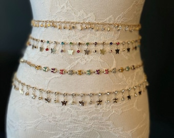 Könnte beinhalten: Ein Set aus fünf goldenen Kettengürteln mit Schmetterlingsanhängern und Kristallverzierungen. Die Gürtel sind auf einem weißen Spitzen-Mannequin übereinander gelegt.