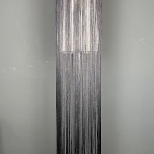May include: A floor-length black fringe garment featuring a high neckline and a sleeveless design. The piece is constructed from numerous thin, vertical black threads that hang straight down from the collar, creating a long, linear silhouette.