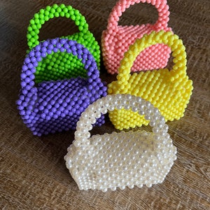 Op de afbeelding: Vijf kralenhandtassen in verschillende kleuren. De handtassen zijn gemaakt van kleine, ronde kralen en hebben een bovenste handvat. De kleuren zijn groen, paars, geel, roze en wit.