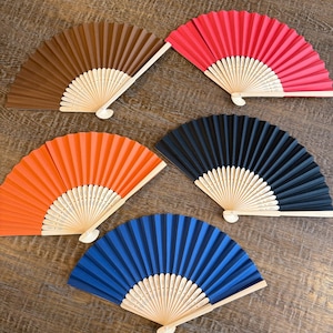 May include: Five folding hand fans with light-coloured wooden ribs and fabric leaves in solid colours: brown, red, orange, black, and blue. Each fan features a pleated design with intricate cut-out patterns on the wooden handle base.