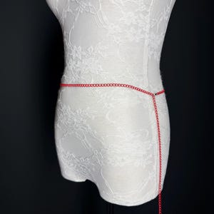 May include: A red chain body accessory displayed on a white lace-covered mannequin. The chain is fine and hangs from the waist, with a longer strand extending downwards. The background is a solid black.