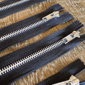 Puede incluir: Cuatro cintas de cremallera negras con dientes de metal plateado. Las cremalleras están dispuestas en fila sobre una superficie de madera.