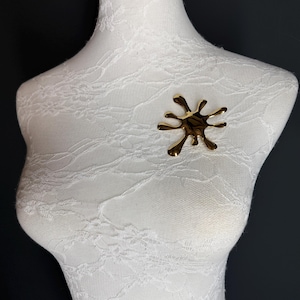 Puede incluir: Un broche abstracto de color dorado en forma de salpicadura o salpicadura. El broche est&aacute; en un maniqu&iacute; blanco.