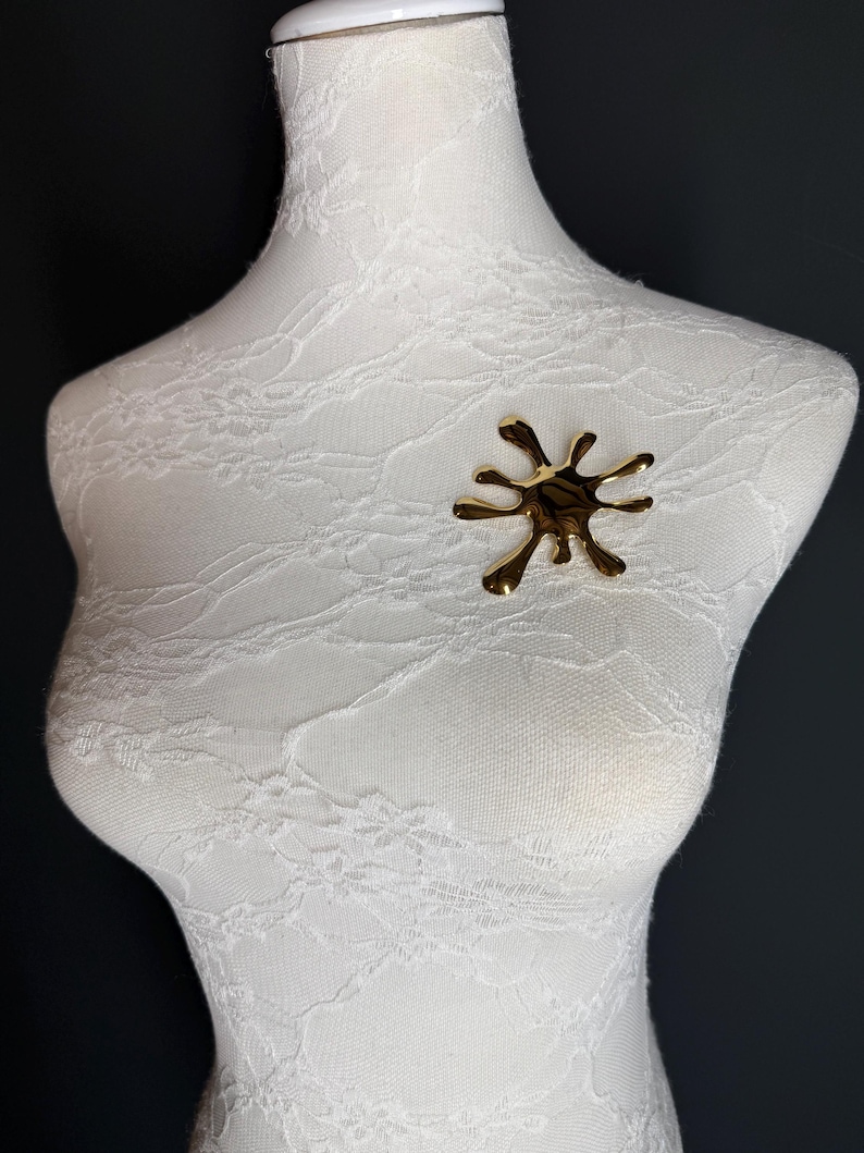 Puede incluir: Un broche abstracto de color dorado en forma de salpicadura o salpicadura. El broche est&aacute; en un maniqu&iacute; blanco.