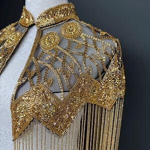 May include: A gold beaded capelet with a high collar and intricate floral and geometric patterns. The capelet features long, dangling fringe and is displayed on a white mannequin. The design is ornate and elegant.