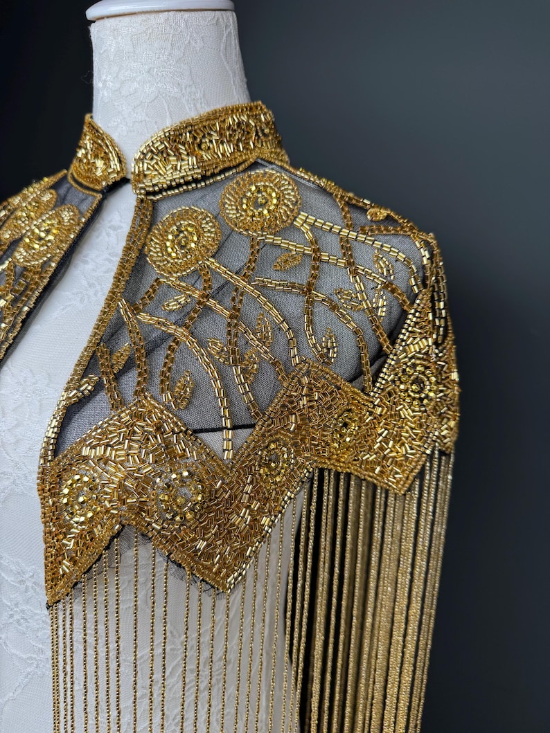 May include: A gold beaded capelet with a high collar and intricate floral and geometric patterns. The capelet features long, dangling fringe and is displayed on a white mannequin. The design is ornate and elegant.