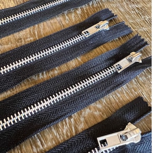 May include: Four black zipper tapes with silver metal teeth and sliders. The zippers are laid out in a row on a wooden surface.
