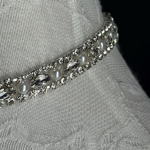 May include: Close-up of a silver-toned choker necklace with faux pearls and clear rhinestones. The necklace is set against a white fabric background, creating a contrast between the shiny jewelry and the textured fabric.