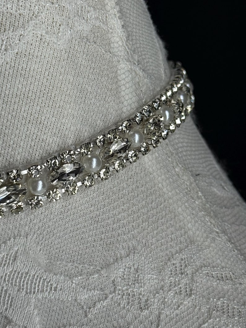 May include: Close-up of a silver-toned choker necklace with faux pearls and clear rhinestones. The necklace is set against a white fabric background, creating a contrast between the shiny jewelry and the textured fabric.