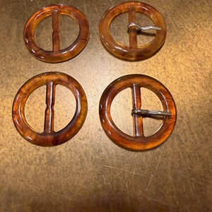 Op de afbeelding: Vier vintage, ronde gespen in schildpadstijl. Elke gesp heeft een cirkelvormig frame met een verticale balk en een metalen pin-sluiting op twee van de gespen. De gespen zijn warmbruin van kleur met een glanzende afwerking.